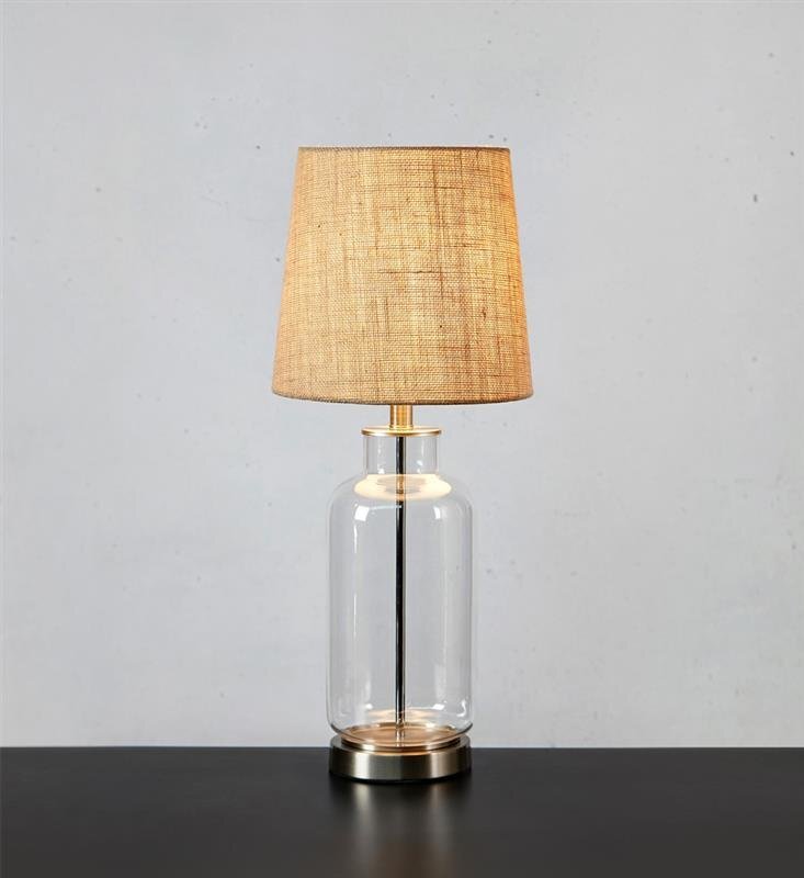 Lampa stołowa z przezroczystą szklaną podstawą i beżowym abażurem, ustawiona na ciemnym blacie, na tle jasnej ściany.