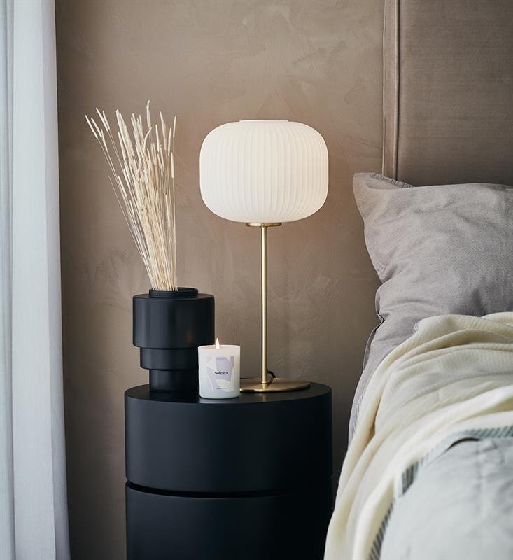 Na zdjęciu znajduje się elegancka lampa stołowa z białym kloszem i złotą podstawą, ustawiona na czarnym stoliku obok świecy i wazonu z suszonymi roślinami. W tle widoczna jest część łóżka z szarą pościelą.
