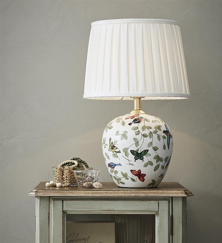 Lampa stołowa z białym abażurem i ozdobną podstawą z motywem roślinnym i motylami stoi na drewnianym stoliku obok szklanej miski z dekoracjami.