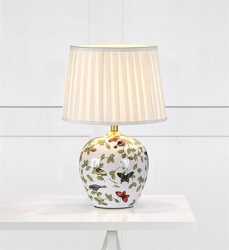 Lampa stołowa z białym abażurem i ozdobną podstawą z motywem liści i ptaków, ustawiona na białym stole na tle jasnej ściany.
