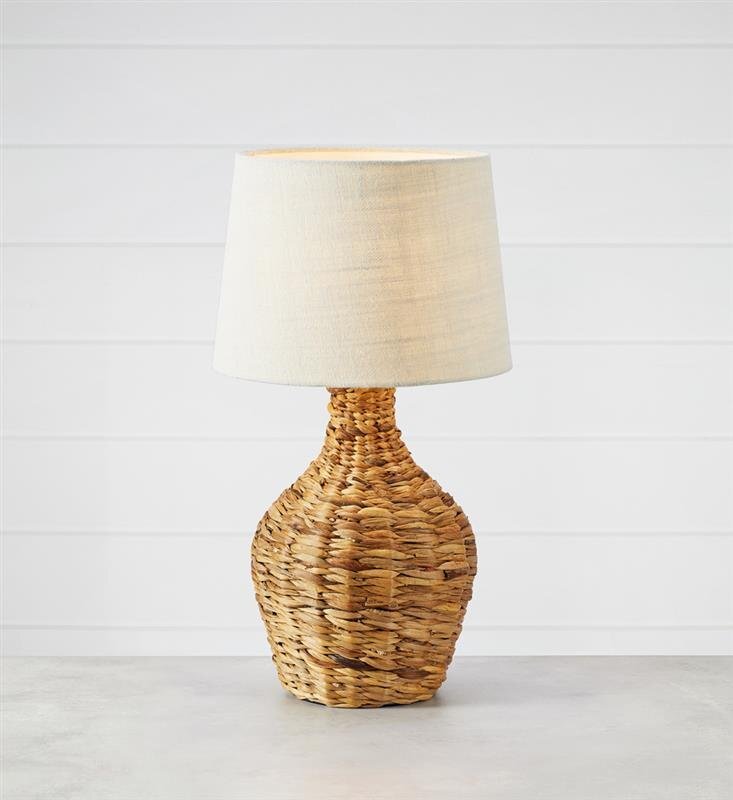 Lampa stołowa z plecioną podstawą w naturalnym kolorze i białym abażurem, ustawiona na jasnym tle.