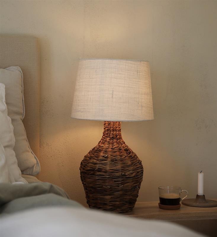 Lampa stołowa z plecioną podstawą i jasnym abażurem stoi na stoliku obok łóżka, obok znajduje się szklanka z napojem i świeca.