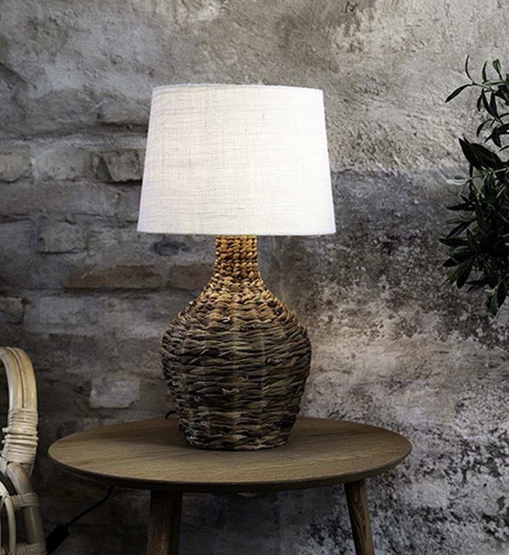 Lampa stołowa z plecioną podstawą i białym abażurem stoi na drewnianym stoliku przed ścianą z surowych cegieł.