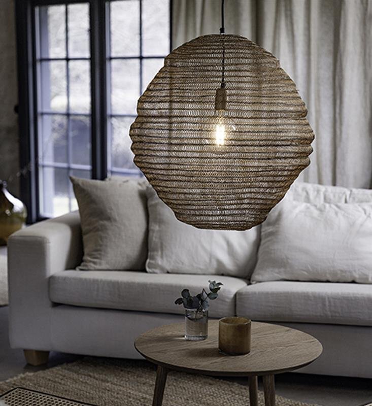 Lampa wisząca o teksturowanym kloszu wisi nad drewnianym stolikiem, na którym znajduje się szklany wazon z rośliną i świeca, w tle widoczna jest sofa z poduszkami oraz zasłony.