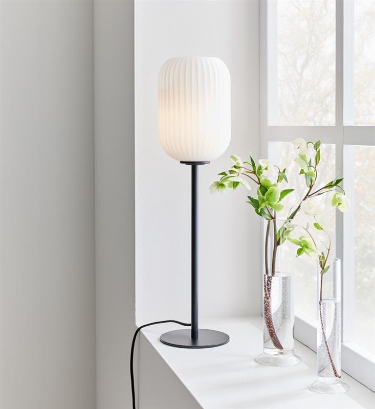 Na parapecie stoi wysoka lampa stołowa z białym, żebrowanym kloszem i czarną podstawą. Obok lampy znajdują się dwie szklane wazony z zielonymi gałązkami.