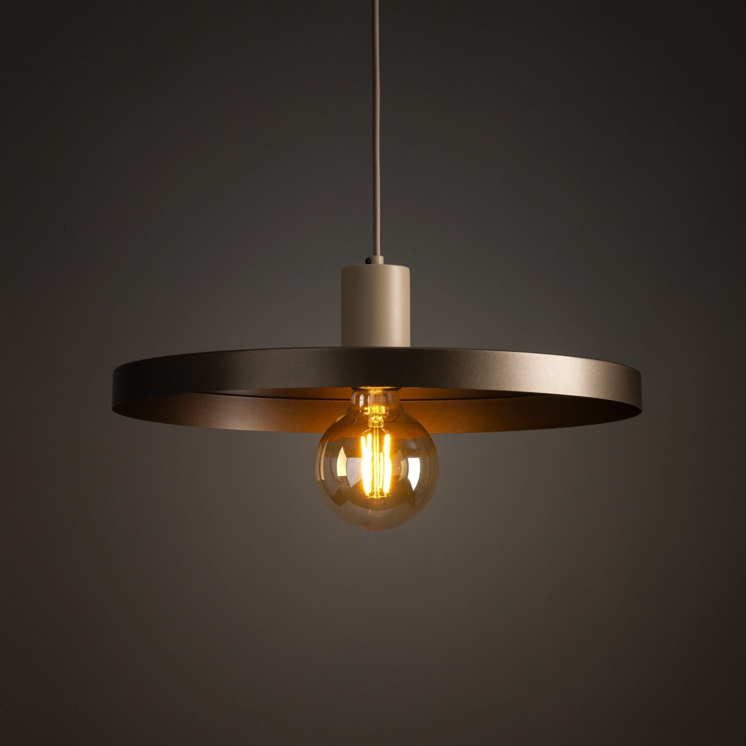 Lampa wisząca z metalowym pierścieniem w kolorze brązowym i widoczną żarówką emitującą ciepłe światło, zawieszona na ciemnym tle.
