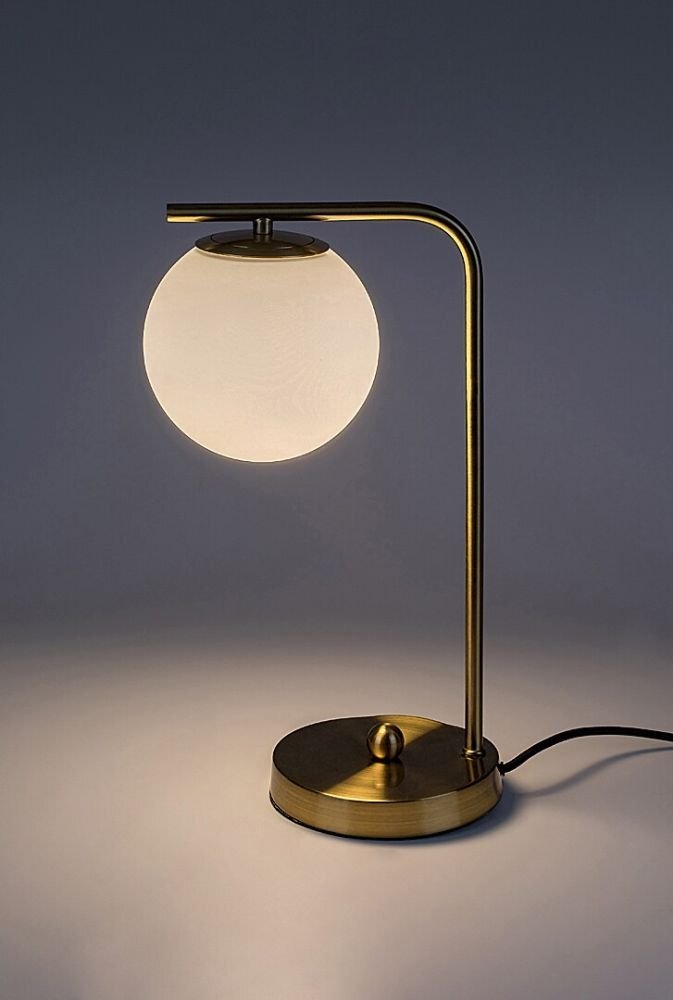 Złota lampa biurkowa z metalowym stelażem i szklanym kloszem w kształcie kuli. Lampa stoi na okrągłej podstawie z widocznym przewodem zasilającym.