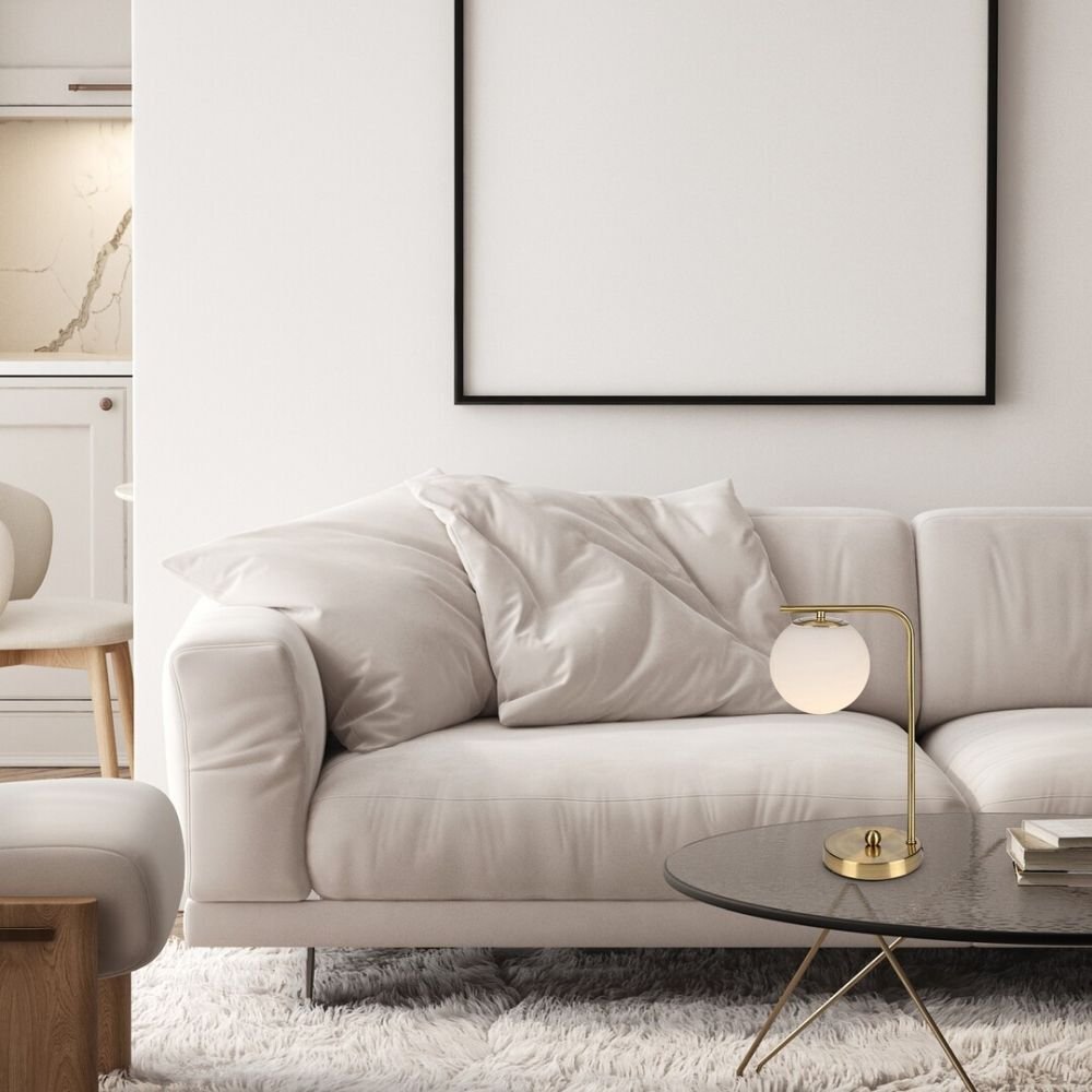 Na zdjęciu znajduje się elegancki salon z białą sofą, na której leżą poduszki, oraz złotą lampą biurkową na okrągłym stoliku. W tle widoczna jest kuchnia z jasnymi szafkami i marmurowym blatem.