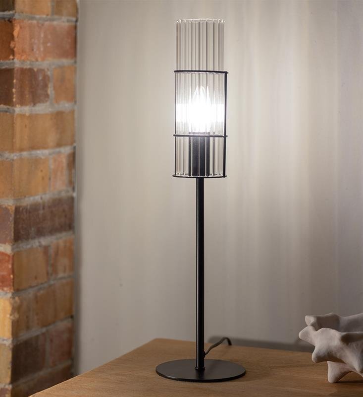 Lampa stołowa z przezroczystym kloszem na czarnym stojaku, umieszczona na drewnianym stole obok ceglanej ściany.