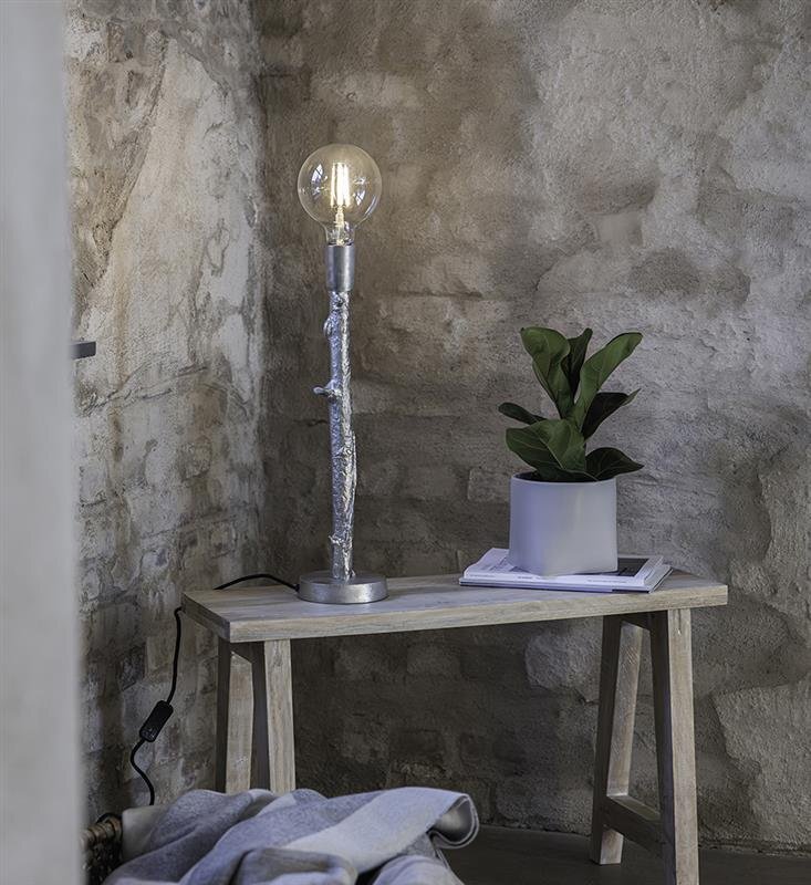 Na drewnianym stoliku stoi lampa stołowa z przezroczystą żarówką oraz doniczka z zieloną rośliną. W tle widoczna jest kamienna ściana.