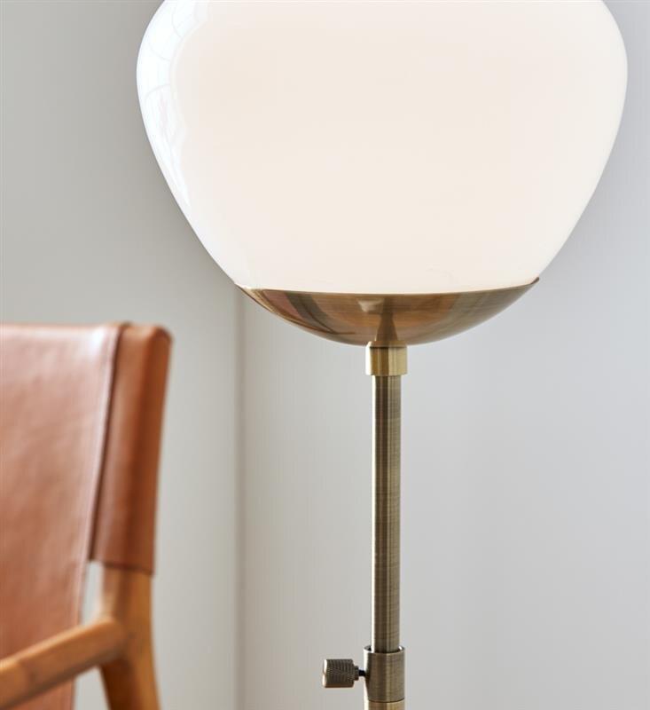 Na obrazie widoczna jest lampa stołowa z białym kloszem i metalową podstawą. Obok lampy znajduje się fragment drewnianego krzesła z brązowym obiciem.