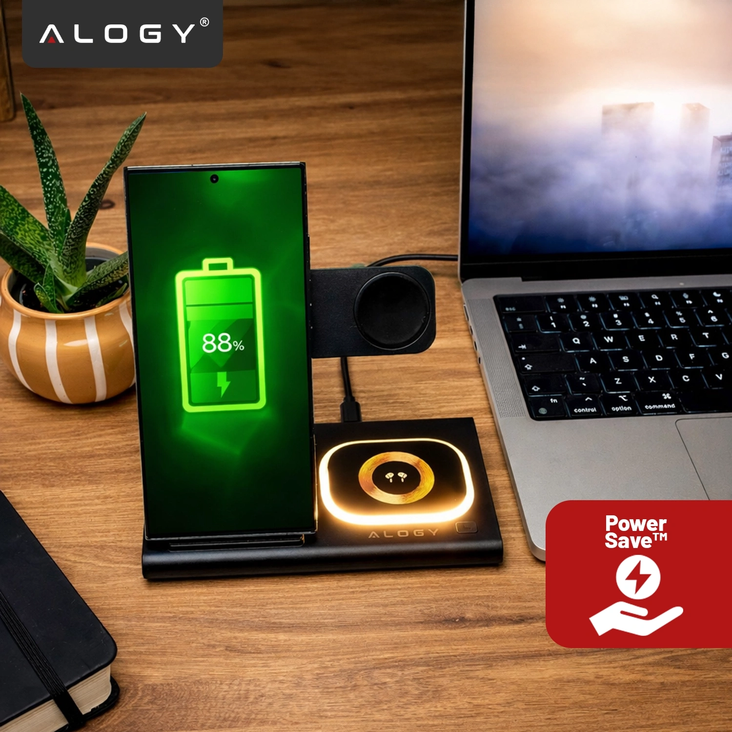 Na drewnianym blacie znajduje się czarna ładowarka indukcyjna z telefonem pokazującym ikonę baterii naładowanej w 88%. Obok ładowarki stoi laptop, a w tle widoczna jest roślina w doniczce oraz logo \'ALOGY\'.