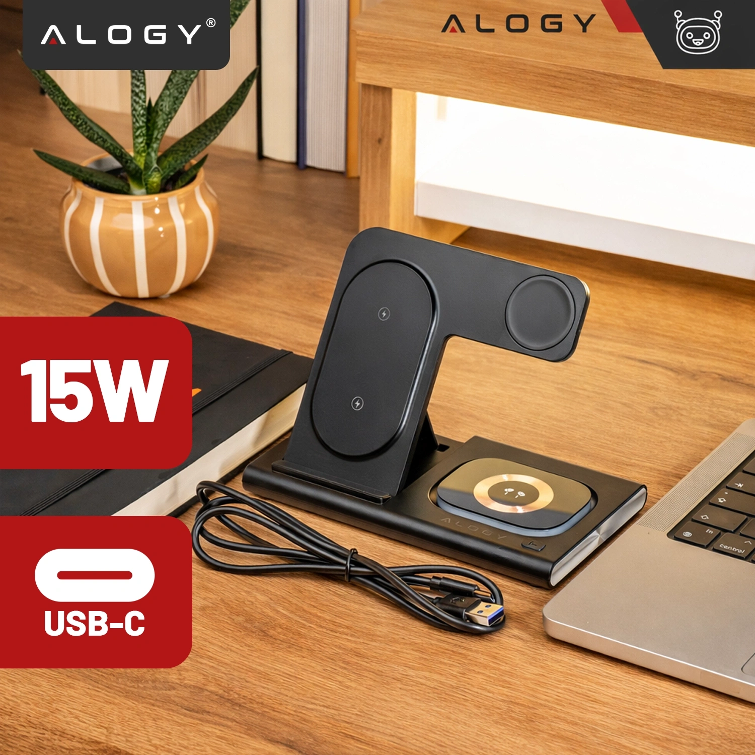 Na drewnianym biurku znajduje się czarna ładowarka indukcyjna z trzema miejscami do ładowania, obok której leży kabel USB-C. W tle widoczna jest doniczka z rośliną oraz książki, a na obrazie znajdują się napisy \'15W\', \'USB-C\' i logo \'ALOGY\'.