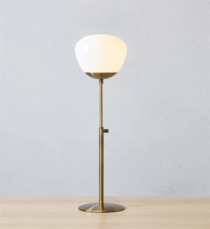 Lampa stołowa z białym kloszem i metalową podstawą w kolorze złotym, ustawiona na drewnianym blacie. Tło jest jednolite, w jasnym odcieniu szarości.