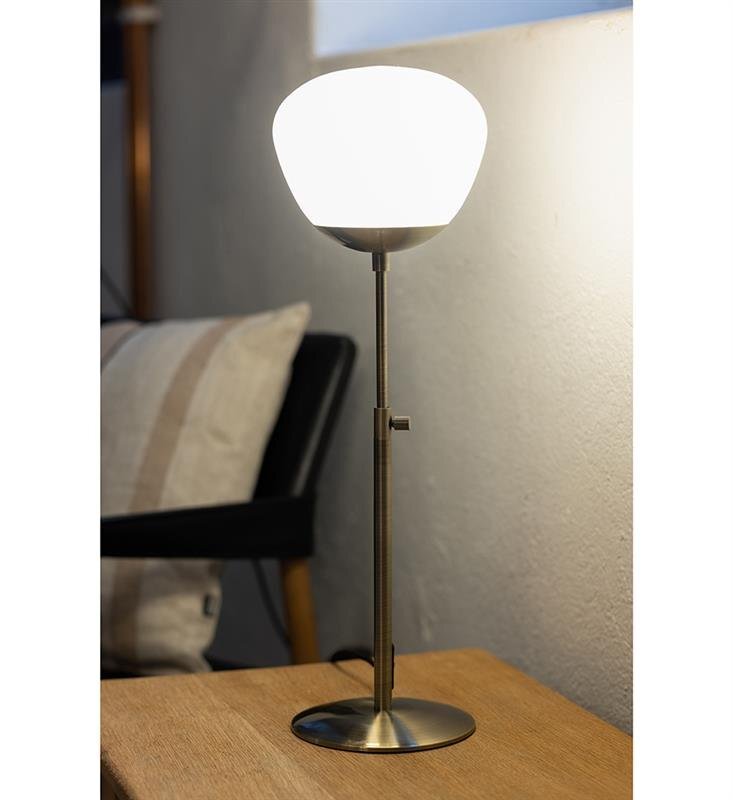 Na zdjęciu widoczna jest lampa stołowa z białym kloszem i metalową podstawą, stojąca na drewnianym stoliku. W tle znajduje się fragment krzesła z poduszką.