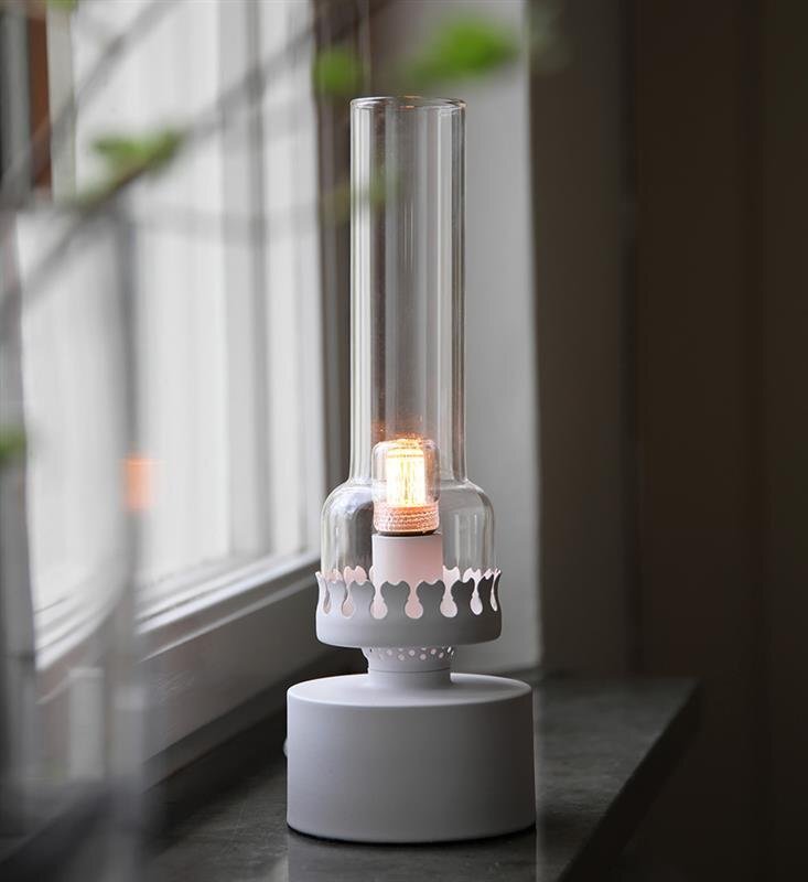 Na obrazie widoczna jest stylowa lampa stołowa z przezroczystym szklanym kloszem i białą podstawą, umieszczona na parapecie przy oknie. Wewnątrz klosza znajduje się żarówka emitująca ciepłe światło.