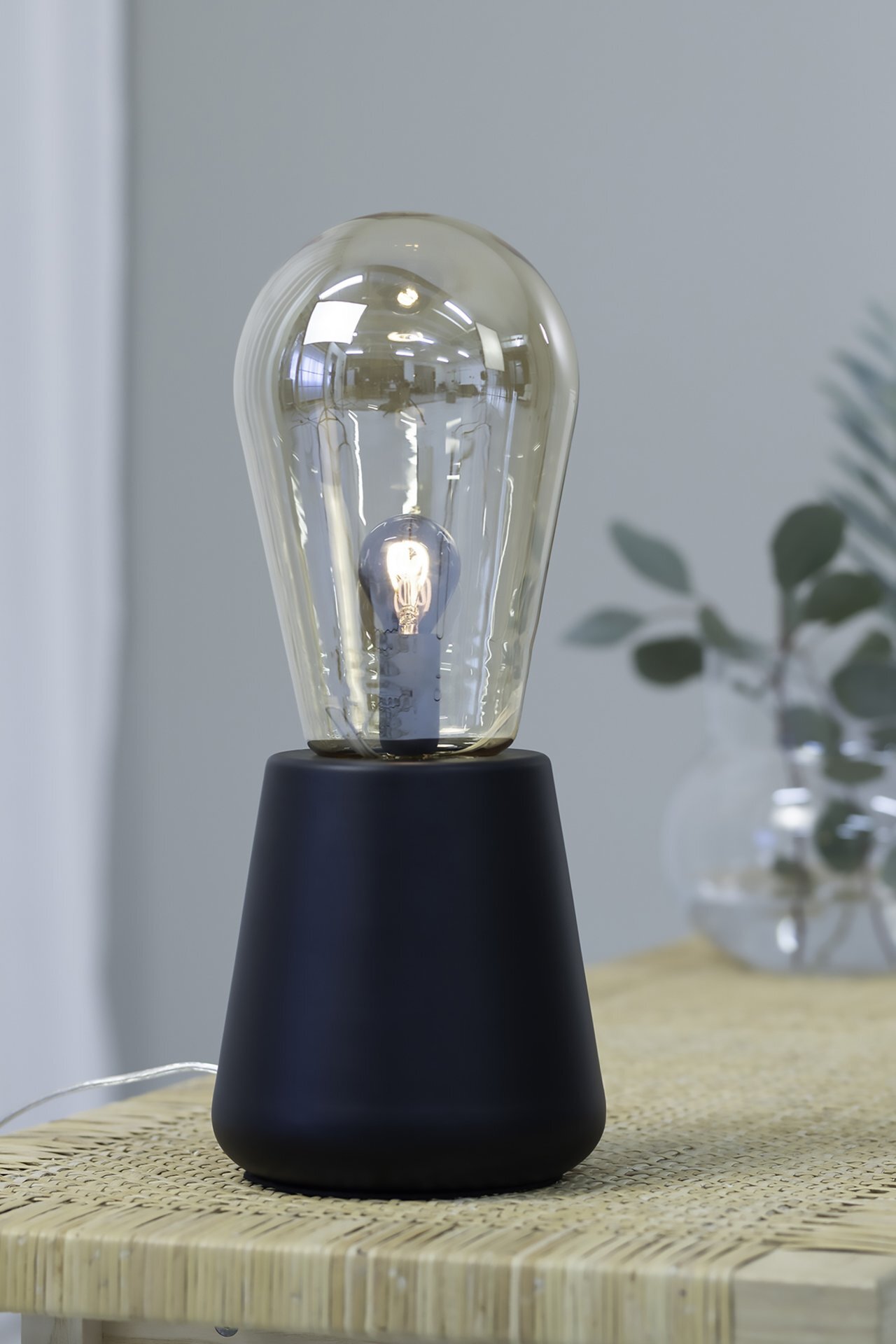 Elegancka lampa stołowa z czarną podstawą i przezroczystym kloszem w kształcie żarówki, ustawiona na drewnianym stole. W tle widoczne są rozmyte elementy dekoracyjne.