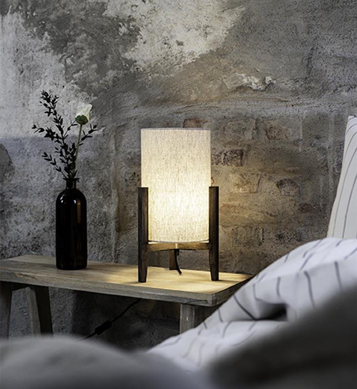 Na drewnianym stoliku stoi lampa stołowa z jasnym abażurem i ciemną podstawą. Obok lampy znajduje się ciemna butelka z pojedynczym kwiatem, a w tle widoczna jest surowa, betonowa ściana.