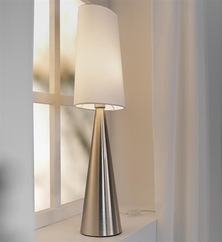 Lampa stołowa o stożkowej podstawie z metalu i białym, cylindrycznym abażurem, ustawiona na parapecie przy oknie. W tle widoczne są zasłony i rozmyty widok za oknem.