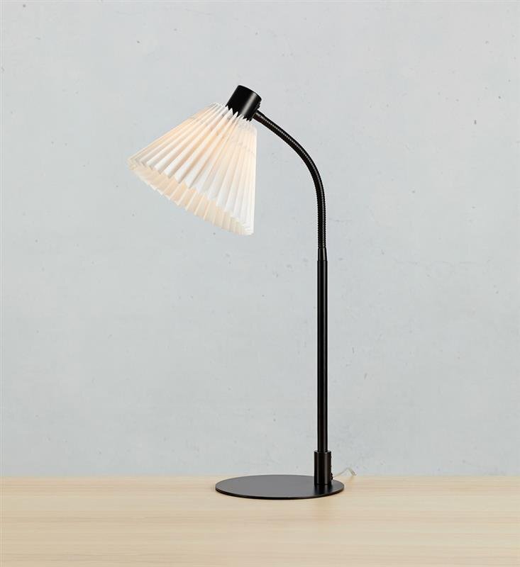Lampa biurkowa z czarną podstawą i elastycznym ramieniem, zakończona białym, plisowanym kloszem. Stoi na drewnianym blacie, na tle jasnej ściany.