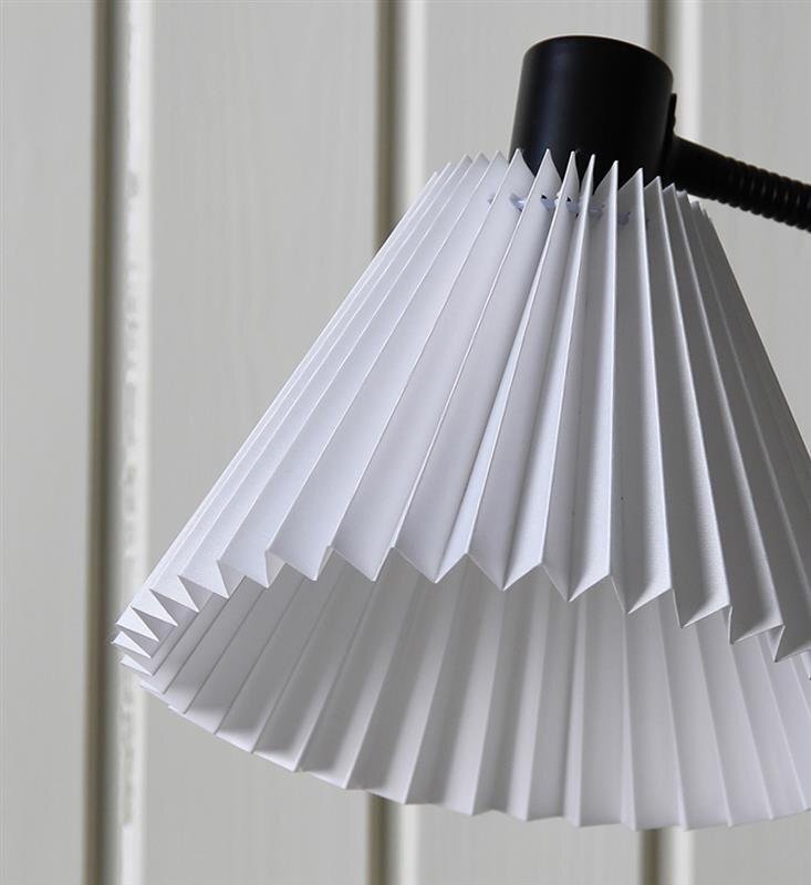 Biała, plisowana osłona lampy biurkowej z czarnym uchwytem, na tle jasnej, drewnianej ściany.