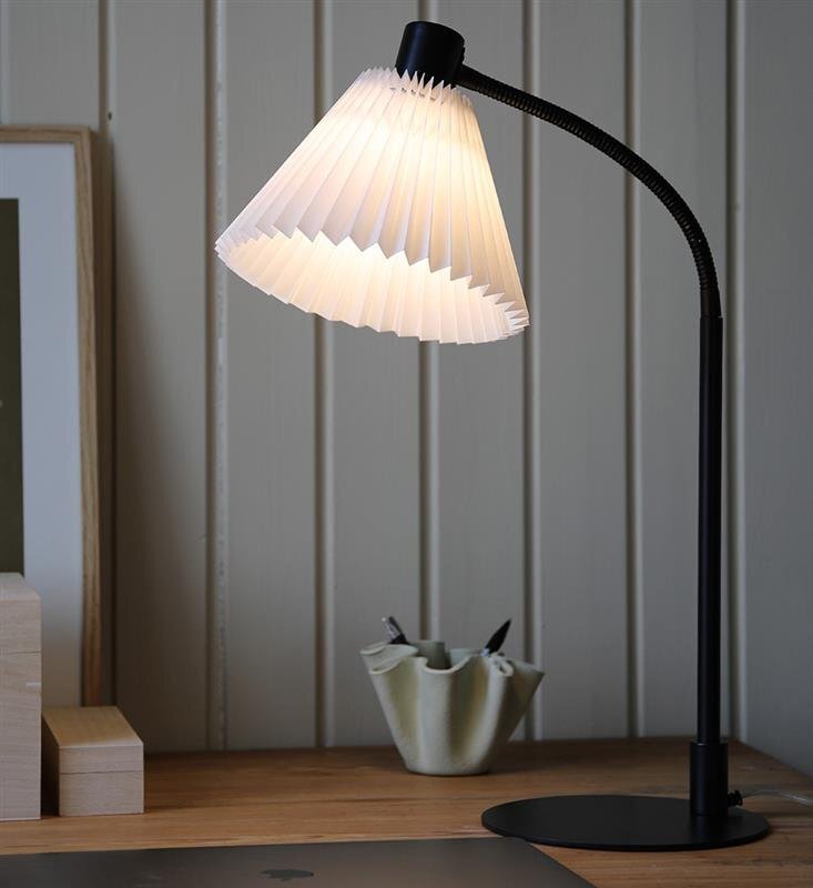 Na drewnianym biurku stoi czarna lampa biurkowa z białym, plisowanym kloszem. W tle widoczne są drewniane panele ścienne i fragment obrazu w ramce.