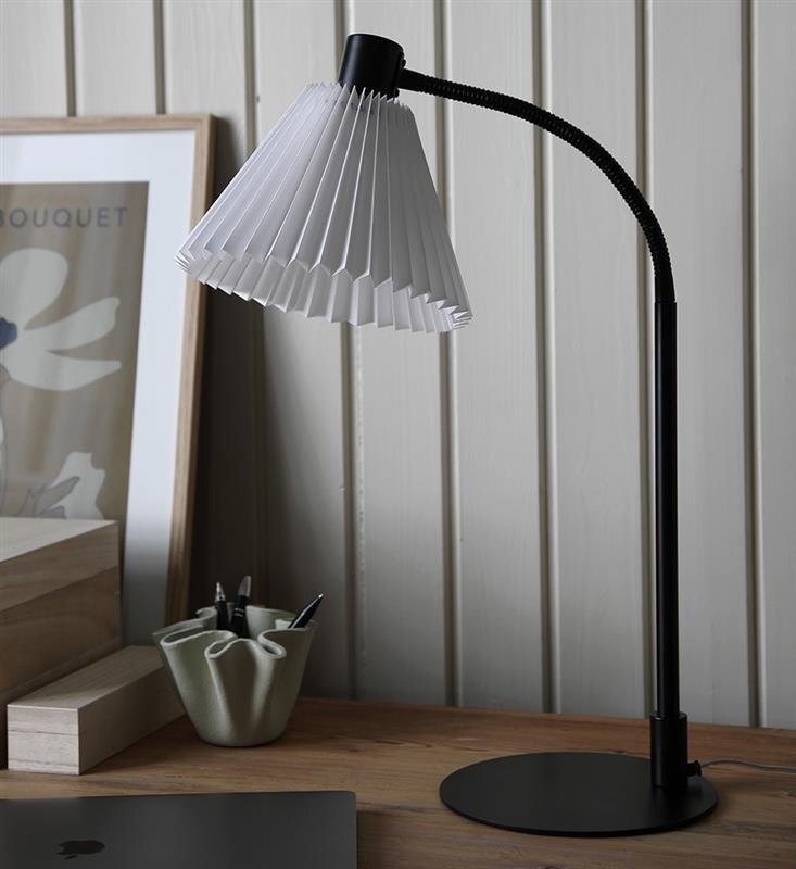 Czarna lampa biurkowa z białym, plisowanym kloszem stoi na drewnianym blacie, obok znajduje się zielony pojemnik z długopisami i oprawiony plakat z napisem \'BOUQUET\'.
