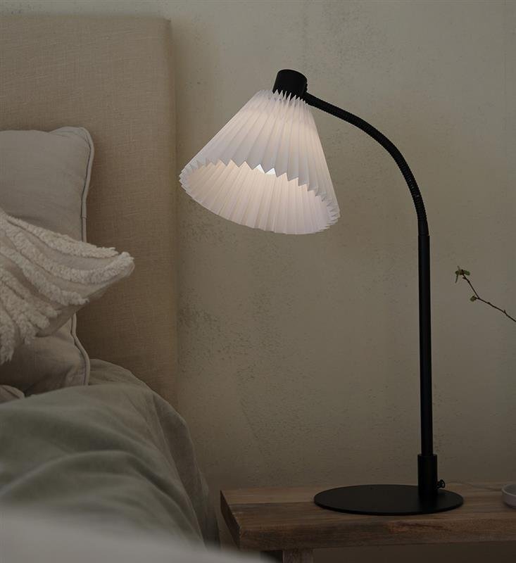 Lampa biurkowa z plisowanym kloszem stoi na drewnianym stoliku obok łóżka z jasną pościelą.