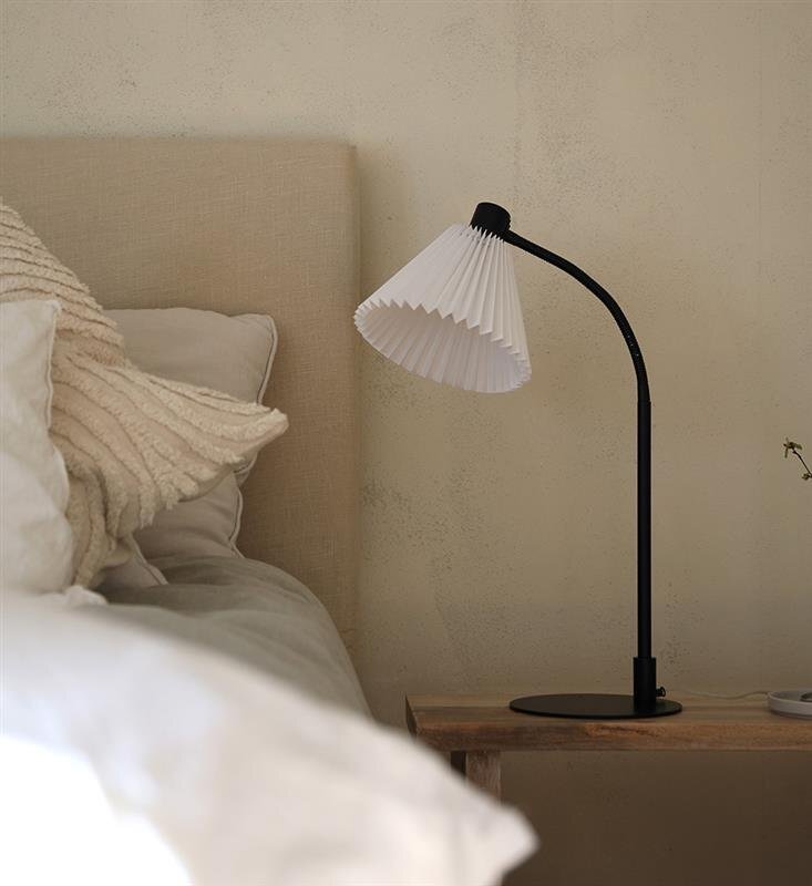 Lampa biurkowa z czarną podstawą i białym, plisowanym kloszem stoi na drewnianym stoliku obok łóżka z jasną pościelą.