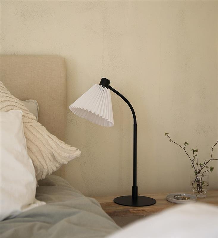 Lampa biurkowa z białym plisowanym kloszem stoi na drewnianym stoliku obok łóżka z jasną pościelą. Obok lampy znajduje się szklany wazon z gałązką oraz mała tacka z biżuterią.