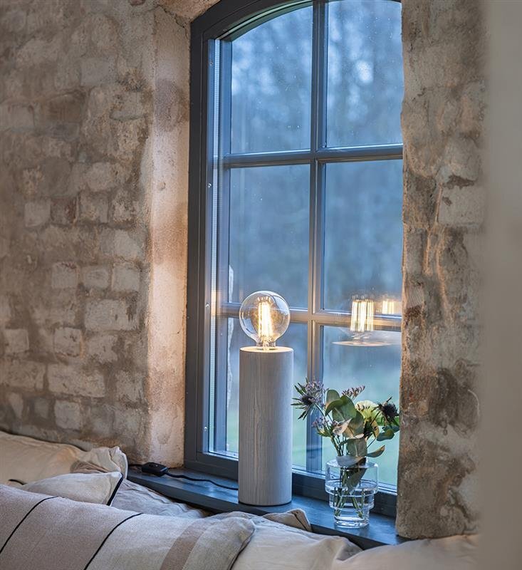 Lampa stołowa nocna z przezroczystym kloszem stoi na parapecie przy oknie, obok wazon z kwiatami. W tle widoczna jest ceglana ściana i zewnętrzny krajobraz za oknem.