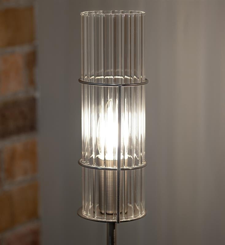 Lampa stołowa z przezroczystym, żebrowanym kloszem, przez który widoczna jest żarówka emitująca jasne światło. W tle widoczna jest ceglana ściana oraz szara powierzchnia.