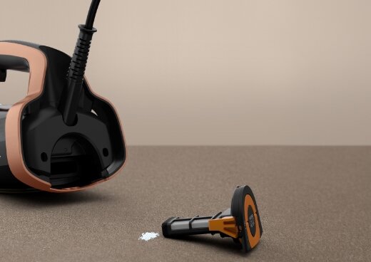 Na obrazie widoczne jest żelazko parowe w kolorze czarnym z elementami miedzianymi oraz wyjęty element, prawdopodobnie filtr, leżący obok. Tło jest jednolite, w kolorze beżowym.