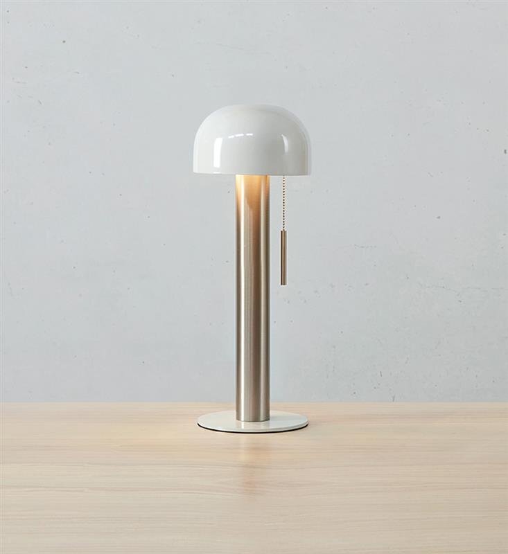 Lampa stołowa z białym kloszem i metalową podstawą stoi na drewnianym blacie, w tle widoczna jest jasna ściana.