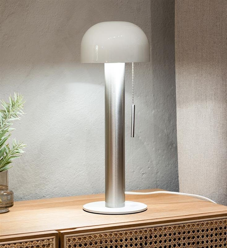 Lampa stołowa z metalową podstawą i białym kloszem, stojąca na drewnianej powierzchni obok zielonej rośliny w doniczce.