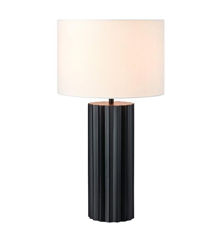 Lampa stołowa z cylindrycznym, czarnym podstawą o żłobionej powierzchni i białym, okrągłym abażurem.
