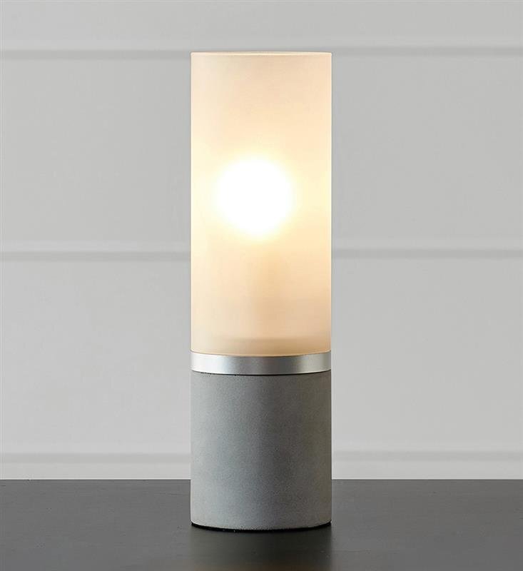 Na obrazie widoczna jest lampa stołowa o cylindrycznym kształcie z jasnym kloszem i ciemną podstawą. Lampa emituje ciepłe, rozproszone światło.