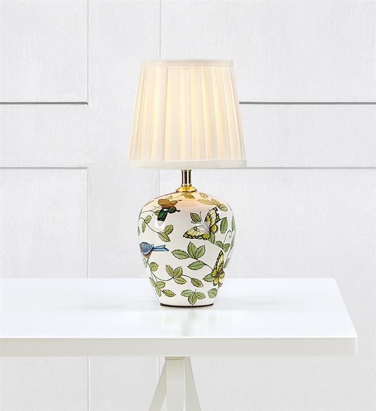 Lampa stołowa z białym abażurem i ozdobną podstawą z motywem roślinnym i motylami, ustawiona na białym stole na tle jasnej ściany.