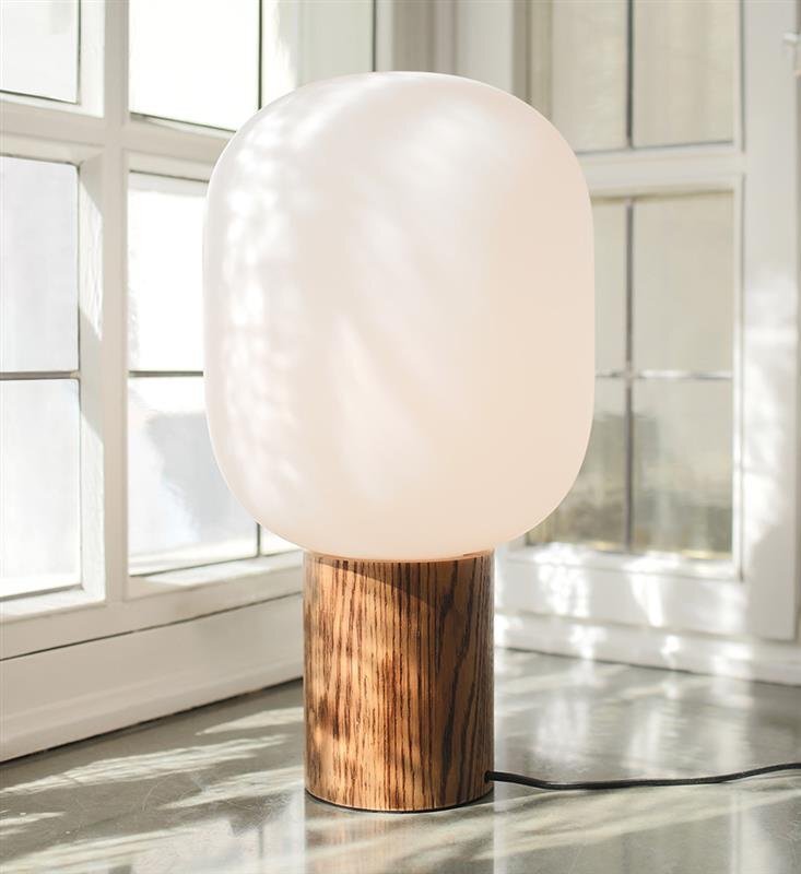 Lampa stołowa z białym, owalnym kloszem i podstawą imitującą drewno, ustawiona na tle okna z kratownicą. Przewód zasilający widoczny na podłodze.