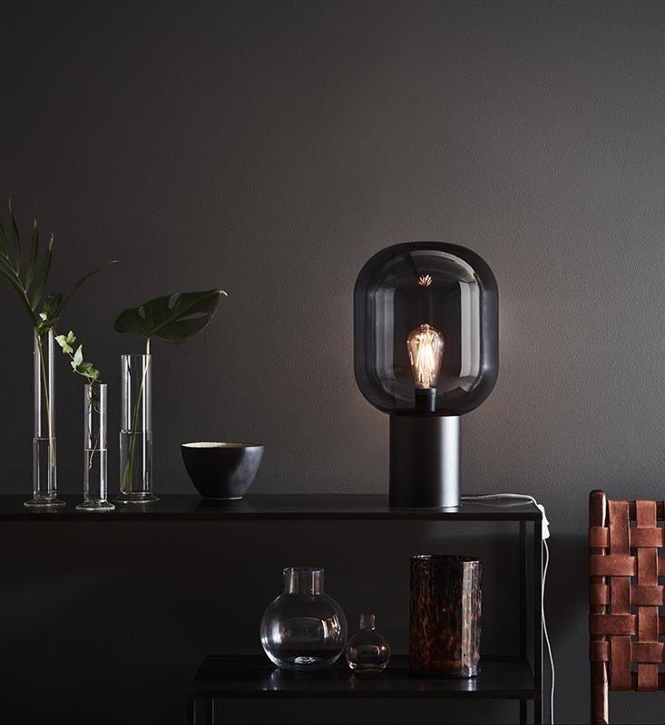 Lampa stołowa z ciemnym kloszem i widoczną żarówką stoi na ciemnym stole obok wazonów z roślinami i dekoracyjnymi przedmiotami.