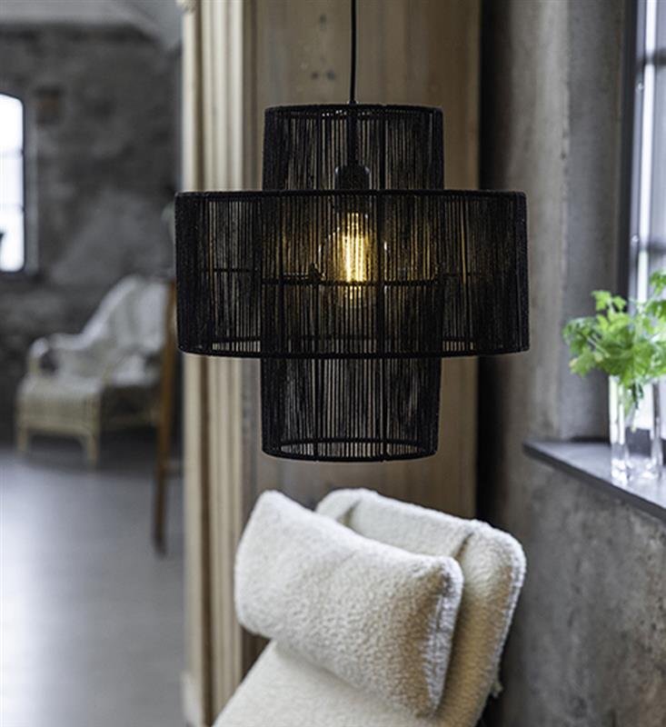 Czarna lampa wisząca z ażurowym kloszem, przez który widać żarówkę, zawieszona w pomieszczeniu z betonowymi ścianami. W tle znajduje się fotel z jasnym obiciem oraz roślina na parapecie.