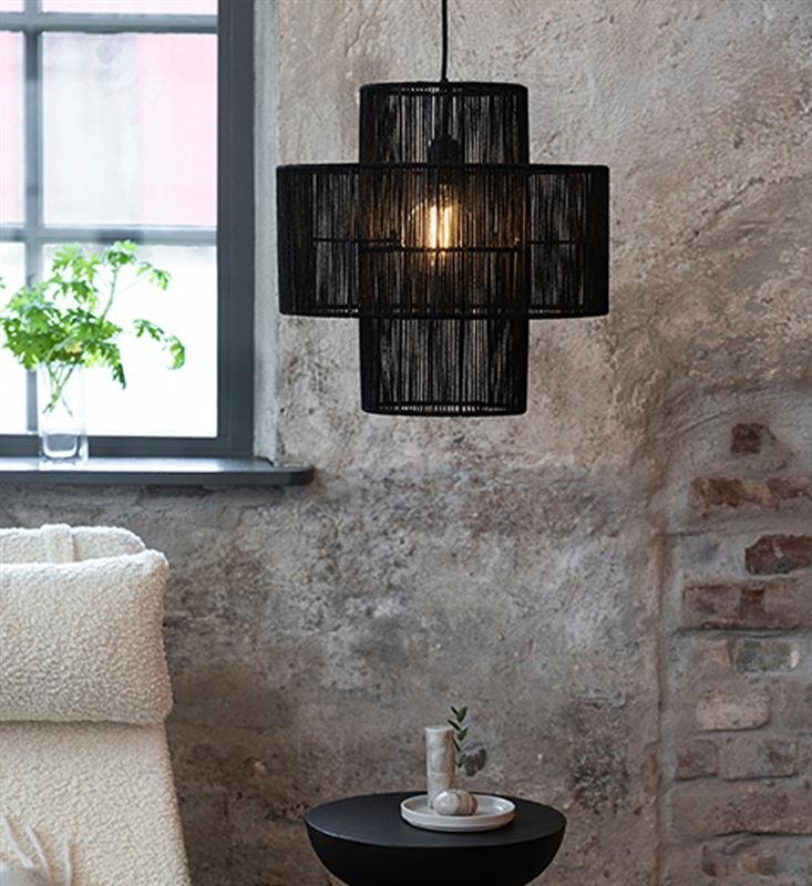Czarna lampa wisząca o geometrycznym kształcie z widoczną żarówką wewnątrz, zawieszona na tle surowej, ceglanej ściany. Obok znajduje się okno z rośliną na parapecie oraz stolik z dekoracją.