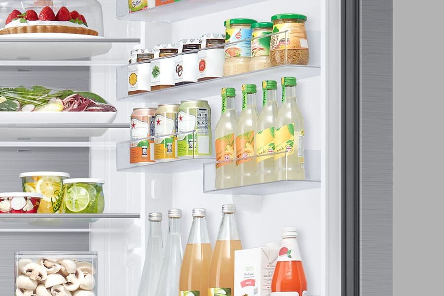 Wnętrze lodówki z półkami wypełnionymi różnorodnymi produktami spożywczymi, w tym napojami w butelkach i puszkach oraz słoikami z przetworami. Na górnej półce widoczny jest talerz z ciastem udekorowanym truskawkami.