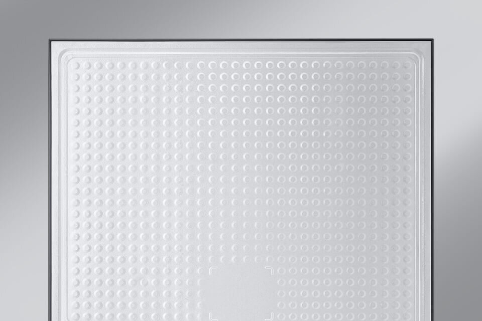 Obraz przedstawia wewnętrzną część lodówki z białą, teksturowaną powierzchnią z wypukłymi kropkami. W tle widoczna jest szara ściana.