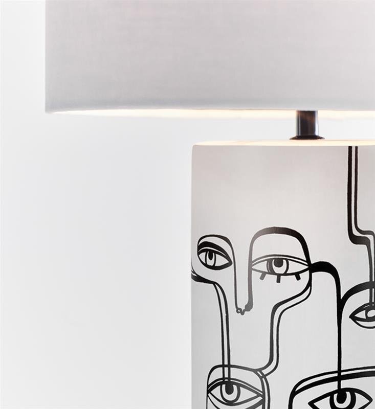 Lampa stołowa z białym abażurem i podstawą ozdobioną czarnymi, abstrakcyjnymi rysunkami twarzy na jasnym tle.