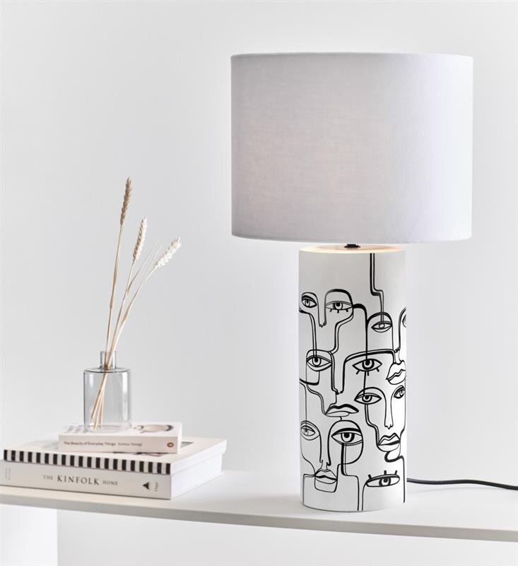 Na obrazie widoczna jest lampa stołowa z białym abażurem i podstawą ozdobioną czarnymi rysunkami twarzy. Obok lampy znajduje się szklany wazon z suszonymi trawami oraz stos książek, w tym jedna z napisem \'THE KINFOLK HOME\'.