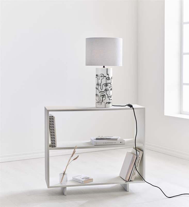 Na białej półce stoi lampa stołowa z białym abażurem i podstawą ozdobioną czarnymi rysunkami twarzy. Obok lampy znajdują się książki i wazon z suchymi gałązkami.