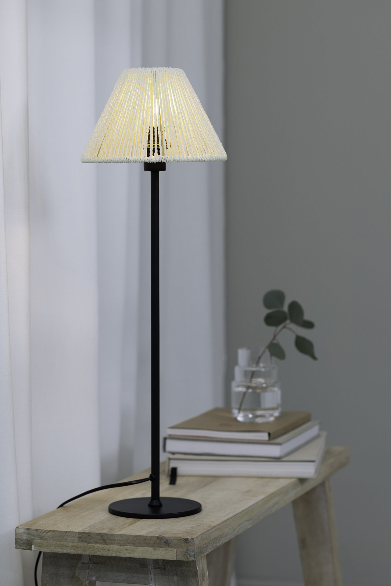 Na drewnianym stoliku stoi wysoka lampa stołowa z czarną podstawą i jasnym, stożkowym abażurem. Obok lampy znajdują się książki i szklany wazon z gałązką.