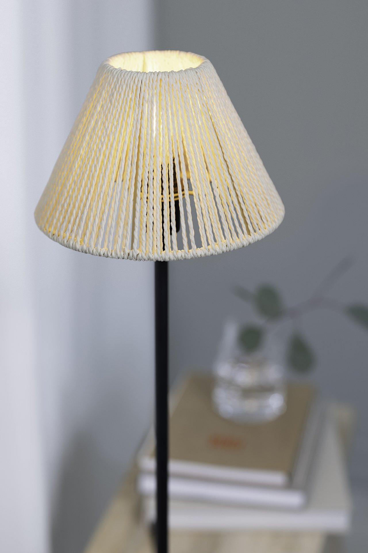 Lampa stołowa z abażurem z jasnego materiału, stojąca na ciemnym trzonku. W tle widoczne są książki i szklanka z gałązką.