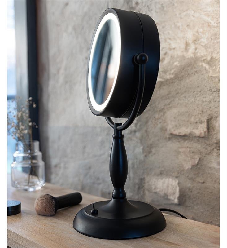 Czarna lampa stołowa z okrągłym lustrem i podświetleniem LED, umieszczona na drewnianym blacie. W tle widoczna jest ściana z surowego kamienia oraz szklany wazon z suszonymi kwiatami.